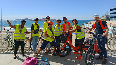 Groupe de cyclistes portant des gilets de sécurité sur le bord de mer à Marseille, avec les îles du Frioul en arrière-plan.